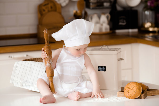 孩子们的厨师。漂亮的小男孩，围着白围裙，戴着厨师帽，手里拿着搅拌器。背景是厨房的内部。烹饪概念。图片下载