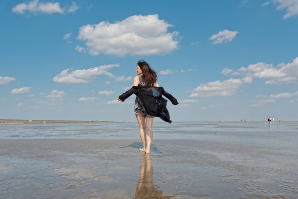 在阳光明媚的夏天，美丽的中国年轻女子黑衣站在泥泞的海滩上，白云和蓝天为背景。无忧无虑的女孩享受她的自由时间。美丽的生活方式。图片下载