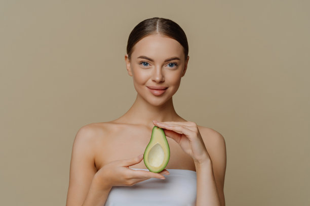 卫生护肤和面部护理理念。室内拍摄满意的女模特抱着一半用浴巾包裹的牛油果准备用美容产品滋养皮肤隔离在米色墙壁图片下载