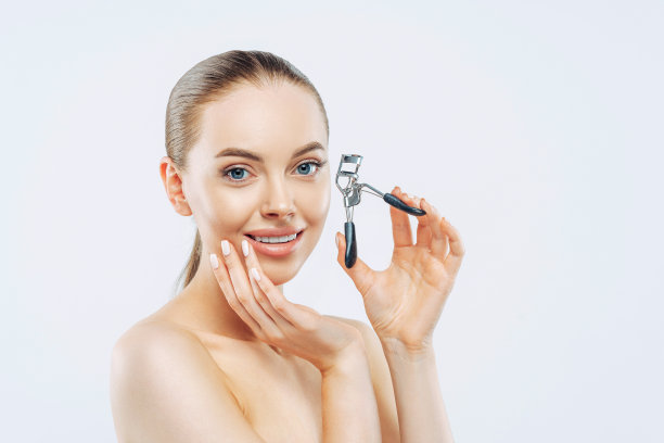 工作室拍摄的年轻黑发女子温柔微笑，使用睫毛夹，光滑柔软的皮肤，站立，有自然的妆容，触摸脸，在白色背景下的姿势。高分辨率图片下载