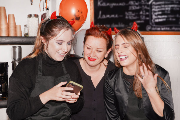 咖啡店的万圣节派对。有趣，大笑，玩得开心，惊讶，扮鬼脸的女服务员穿着万圣节服装，看着手机，智能手机。万圣节面包店。咖啡店，酒吧，柜台，咖啡馆概念。青年企业家的概念图片下载