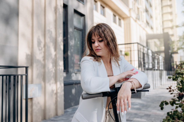 美丽的年轻女子穿着白色西装站在她的电动机车附近的现代建筑在城市和望向别处图片下载
