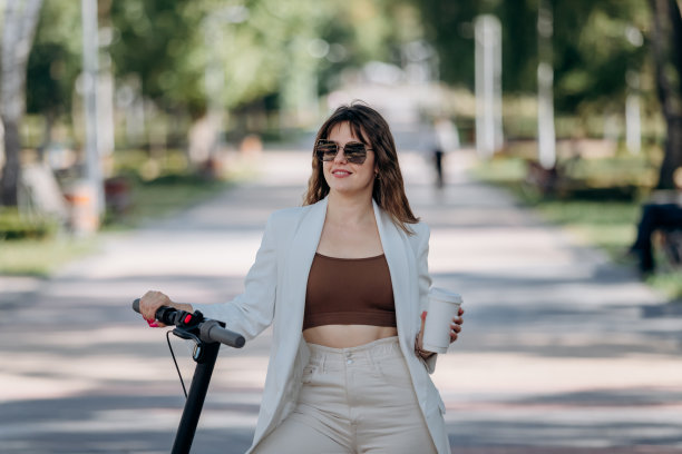 美丽的年轻女子穿着白色西装，戴着墨镜，站在她的电动滑板车在城市公园里喝着咖啡图片下载
