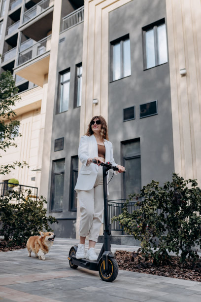 美丽的年轻女子戴着墨镜，穿着白色西装，和柯基狗一起骑着电动车，在城市的现代建筑附近，看着别处图片下载