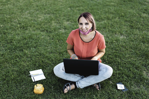 在冠状病毒爆发期间，40岁的女性用笔记本电脑在户外工作-微笑的女性企业家坐在草坪上，一边喝果汁，一边吃早餐-远程工作理念融入大自然图片下载