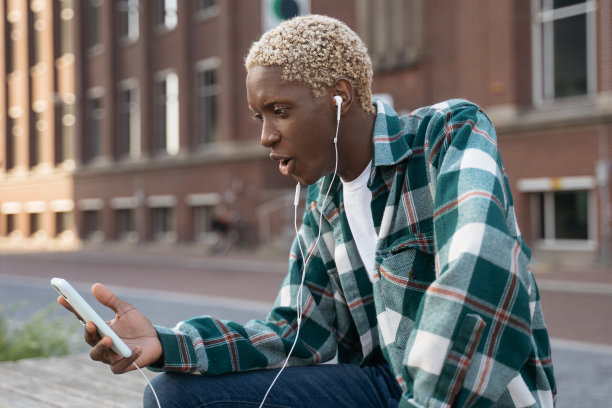 兴奋的非洲男子拿着智能手机在街上玩手机游戏。发型时尚的潮人，穿着休闲的衣服，使用手机应用程序在线购物图片下载