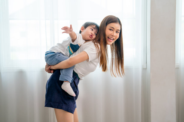 亚洲小男孩骑在妈妈的背上，享受在他们的房子的卧室里一起玩耍，白天的光线透过白色的窗帘和玻璃窗户。图片下载