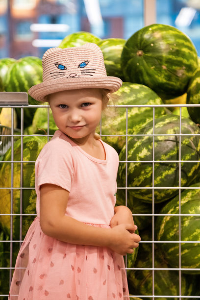 肖像小女孩站在杂货店食品水果店，深思熟虑，看着相机的西瓜背景。可爱可爱的孩子买新鲜水果。选择健康的食品理念。本空间图片下载