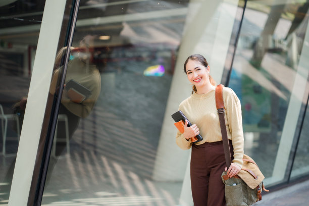 一位拿着平板电脑和肩包的亚洲妇女走在教育理念大楼旁边图片下载