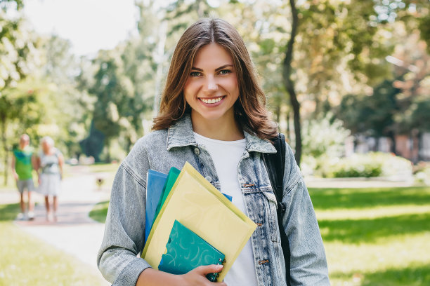 年轻的女学生在公园里微笑。可爱的女学生手里拿着文件夹和笔记本。学习,教育理念图片下载