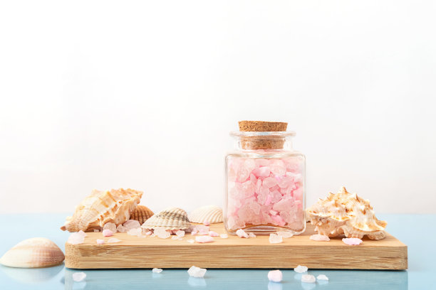 粉红色的海盐装在透明的玻璃瓶里，贝壳放在浅米色的木架子上。环保护理有机产品。美容治疗，水疗理念。副本的空间。图片下载