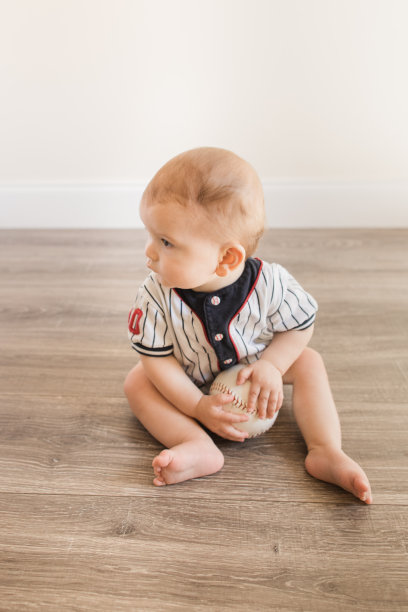 7个月大的小男孩穿着棒球服，一边玩垒球，一边学习一种新的发展技能，在家里的木地板上自己坐起来图片下载