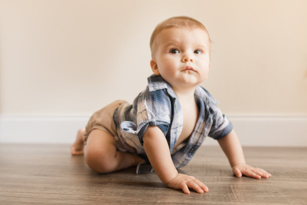 2022年秋季，患有多指畸形的7个月大男婴穿着中性颜色可重复使用的布尿布和蓝色格纹衬衫在木地板上爬行，学习一种新的发育技能图片下载