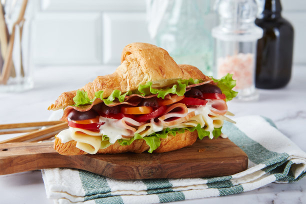 羊角面包三明治配芝士、火腿、生菜、西红柿、橄榄和美味的酱汁，铺在木板上。图片下载