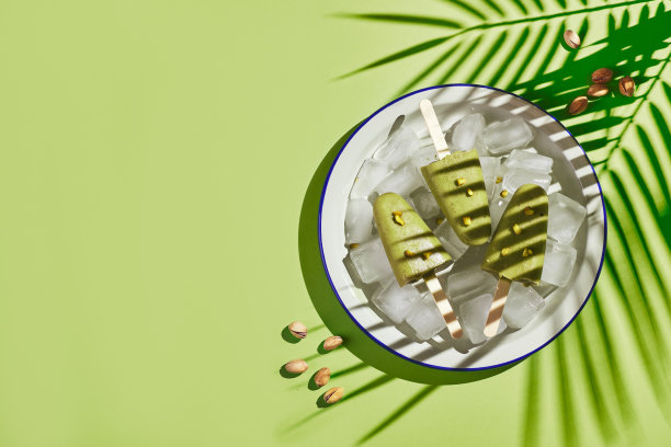 冰冻自制开心果冰棒在碗冰在绿色的背景与棕榈叶的阴影在刺眼的光。清爽的冰棒，冰冻的绿汁棒。俯视图，复制空间图片下载