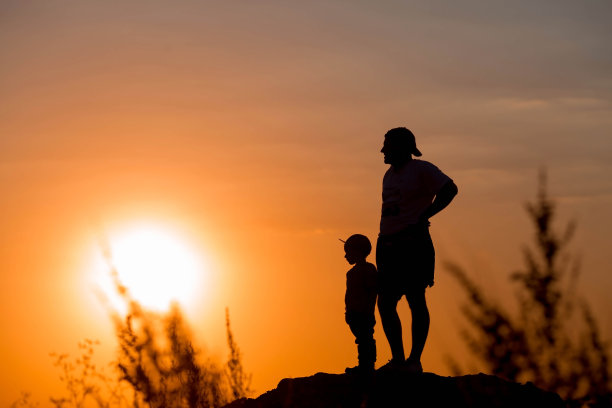 日落时分，父子俩在公园里玩耍。人们在大自然中获得乐趣。橘黄色背景上人们的黑色轮廓。一个友好家庭的概念和暑假以及父亲节。图片下载