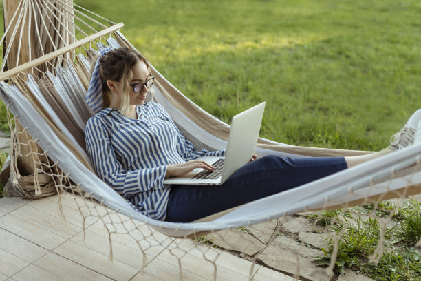 年轻美丽的女人躺在吊床上，带着眼镜的笔记本电脑在外面的家里图片下载