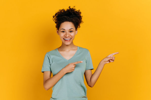 年轻的卷发非洲裔美国女性穿着基本款t恤，表情积极，站在孤立的橙色背景上，看着镜头，手指指向一边，微笑图片下载