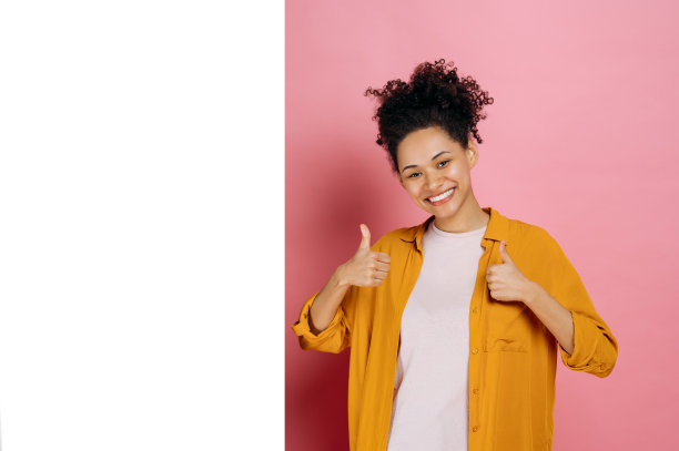 实物模型。兴奋的卷发非洲裔美国女孩，穿着休闲服装，竖起大拇指，站在空白的地方做广告或展示，在粉红色的背景上，看着镜头，微笑图片下载
