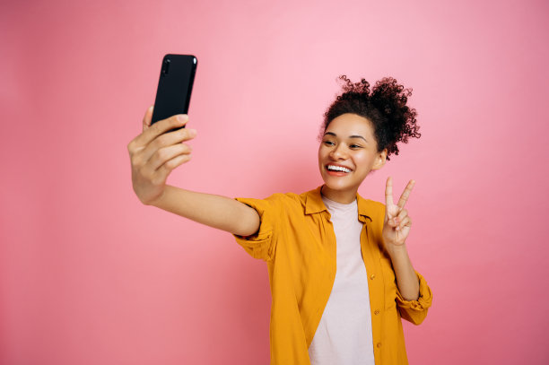 激动的现代非洲裔美国年轻女性，博主，穿着休闲时尚的衣服，用手机自拍，对着相机微笑，展示和平符号，在孤立的粉红色背景上摆姿势，微笑图片下载