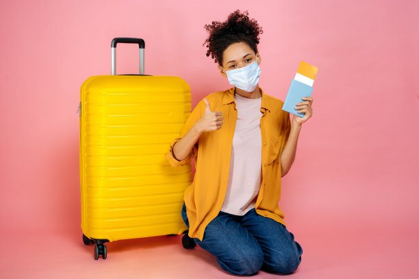 全长照片快乐的非洲裔美国女孩与医疗口罩在她的脸上，持有护照和旅行机票，坐在附近的大黄色手提箱孤立的粉红色背景，看着相机图片下载