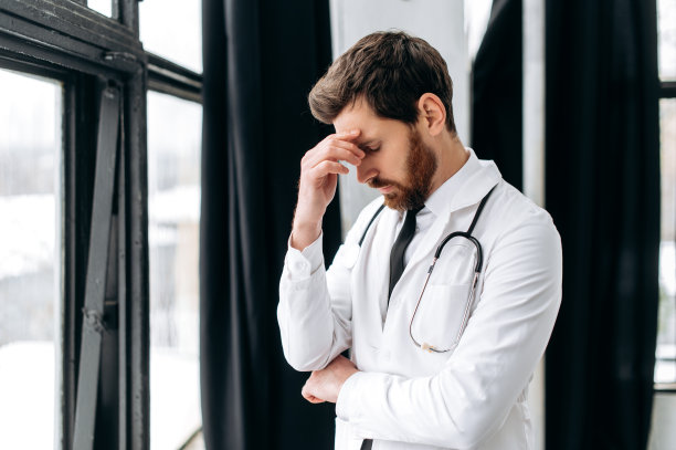 照片上是一位劳累过度的男性全科医生，他身穿白色的医疗制服，肩上扛着听诊器，站在医院靠窗的地方，低着头，需要休息图片下载