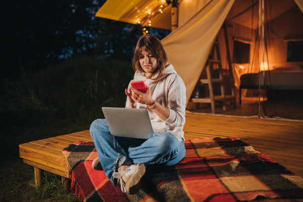 快乐女人自由职业者，夏夜在舒适的露营帐篷里用手机和笔记本电脑工作。用于户外度假和度假的豪华露营帐篷。生活方式的概念图片下载