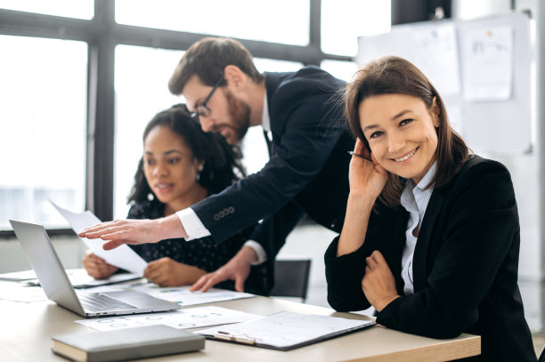 美丽自信的白人女性穿着正装，公司ceo、财务经理，与多种族同事坐在办公室的办公桌前，看着镜头，友好地微笑图片下载