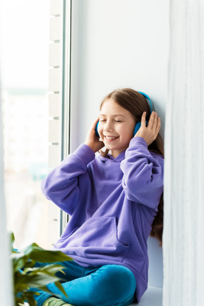 可爱的小女孩戴着无线耳机坐在窗边。孩子在手机应用上听喜欢的音乐并微笑。在家休闲图片下载