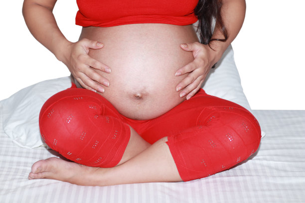 年轻的孕妇坐在床上，手放在肚子上图片下载