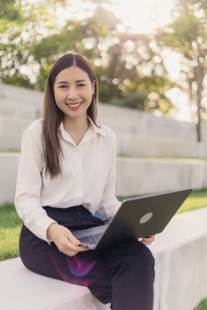 亚洲女商人微笑着看着镜头，手里拿着笔记本电脑，坐在公园里工作。图片下载