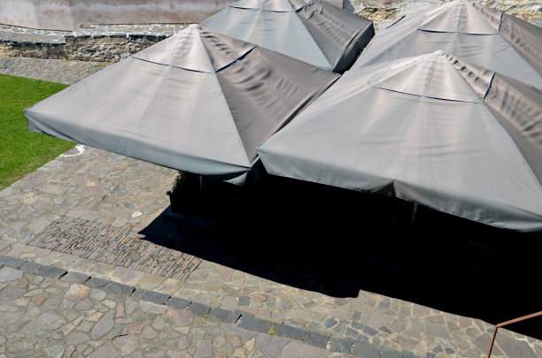 餐馆的石砌露台上有四把闪闪发光的灰色织物遮阳伞，公园花园里的咖啡馆里有金字塔状的防水帆布帐篷，可以遮挡阳光。广场，后院，彼此紧挨着图片下载