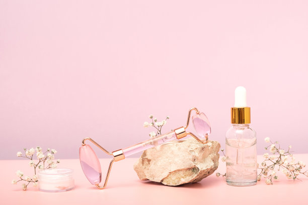 粉嫩玉辊面部按摩水粉和化妆瓶在石头上粉红色的背景与白色的花朵的石膏。家庭水疗护理理念，美丽青春，抗衰老。图片下载