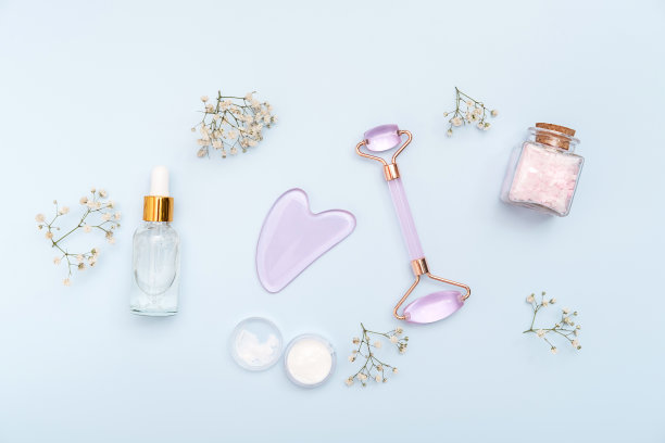 粉色石英石滚筒面部按摩粉彩粉彩蓝色的背景与玻璃化妆瓶和白花。家庭水疗护理理念，美丽青春，抗衰老。前视图。平的。图片下载