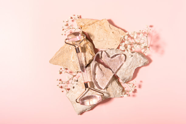 粉色石英石滚筒面部按摩粉彩粉彩背景石头和白花。家庭水疗护理理念，美丽青春，抗衰老。前视图。平的。图片下载