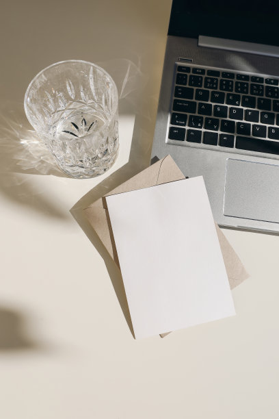 优雅的工作空间组成，打开笔记本电脑。贺卡，请柬模型。一杯水，在夏日的阳光下畅饮。长长的影子覆盖。米色表格背景。经营理念。垂直flatlay图片下载