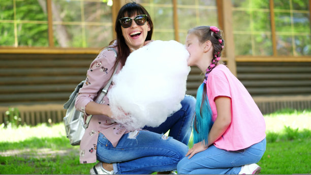 一个深色头发的女人，戴着墨镜，和她的女儿，8岁的女孩，留着蓝色的辫子，在夏天的城市公园里，吃着甜棉花糖，棉花糖，笑着，玩得很开心图片下载