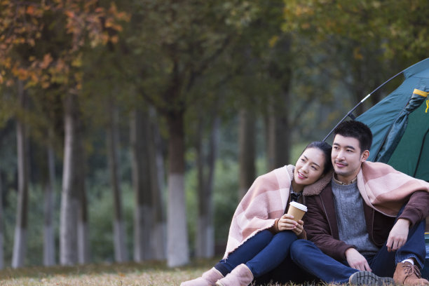 在秋天的丛林草地上，一对年轻夫妇依偎在他们的露营帐篷前，围着同样的披肩喝着咖啡——照片图片下载
