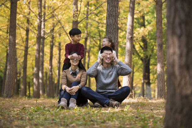 秋天的森林里，年轻的中国父母坐在树下和孩子们玩耍，孩子们用手挡住父母的眼睛——照片图片下载