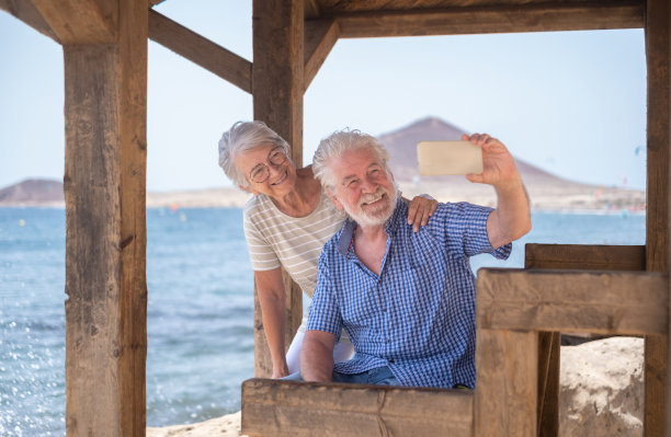 快乐的高加索老年夫妇坐在阴凉凉亭前的大海使用手机自拍-老年人退休享受在一个有风的阳光的日子海上度假图片下载