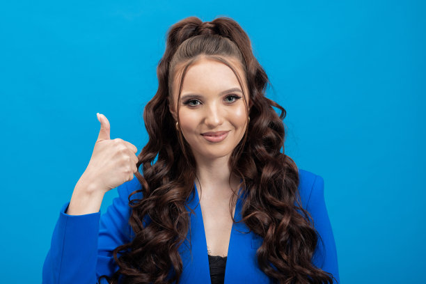 年轻女子微笑，快乐，满意褐发在时尚的夹克显示拇指举的手势，孤立在蓝色背景的工作室肖像人的生活方式概念图片下载