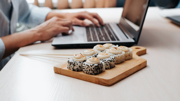 特写的男人在家庭办公室的笔记本电脑上工作，而坐在餐桌上吃传统的日本料理，侧视图。有选择地在木板上烤热寿司图片下载