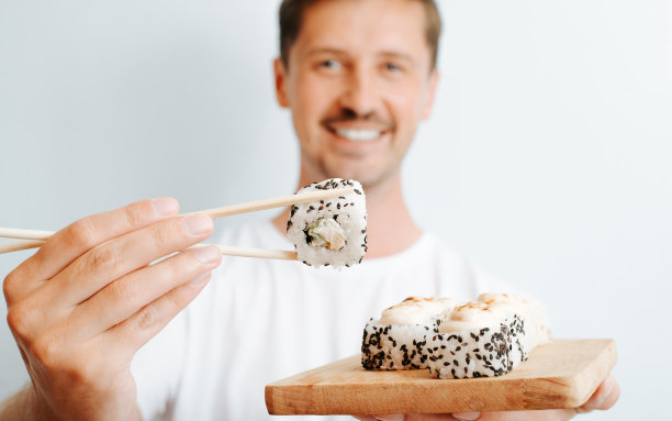 寿司是日本料理的传统食物。一个欢快微笑的男人用筷子夹着米饭卷并向镜头展示。选择性聚焦前景图片下载