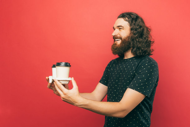 蓄着胡子的快递员穿着一件有黄色图案的黑色t恤，站在红色的墙上，兴高采烈地拿着两个盛着热饮的纸杯，微笑着看着别人图片下载