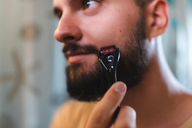 一个秃顶的年轻人在浴室的镜子前用dermaroller修剪他的胡子的特写图片下载