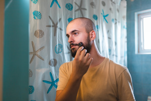 一个秃顶的年轻人在浴室的镜子前用dermaroller修剪他的胡子的特写图片下载