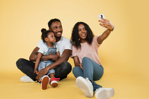 年轻的非洲父母和他们的女儿坐在地板上，拥抱着用智能手机自拍图片下载