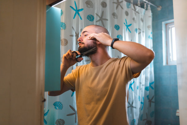 一个秃顶的年轻人在浴室的镜子前用剪刀修剪他的胡子图片下载