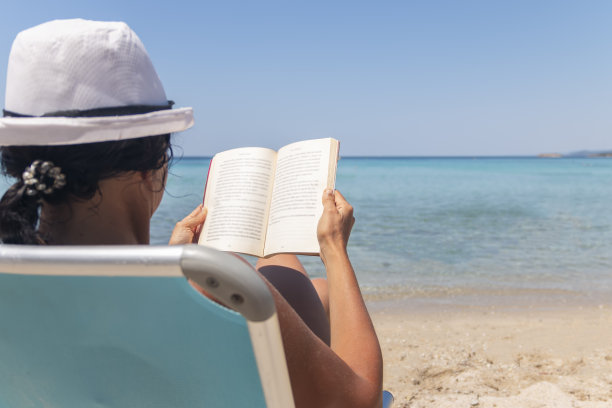 一个幸福的女人坐在躺椅上，在海滩上看书的肖像图片下载