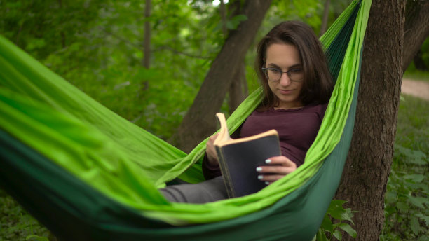 一个年轻的女人躺在吊床上看书。一个戴眼镜的女孩在大自然中进行自我发展。图片下载